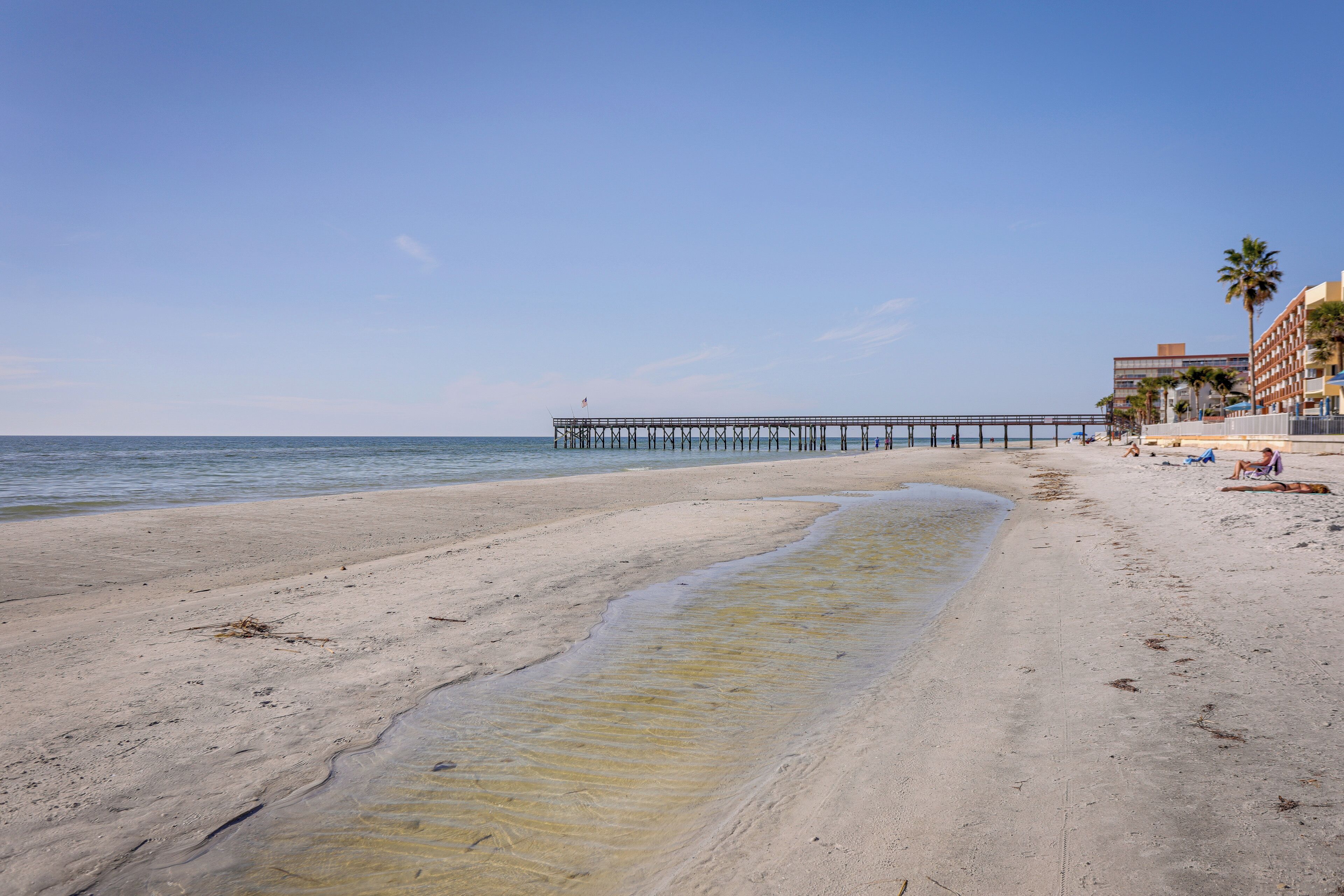 A beach with a pier extending into the water, sandy shore, and buildings in the background.