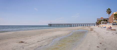 A beach with a pier extending into the water, sandy shore, and buildings in the background.