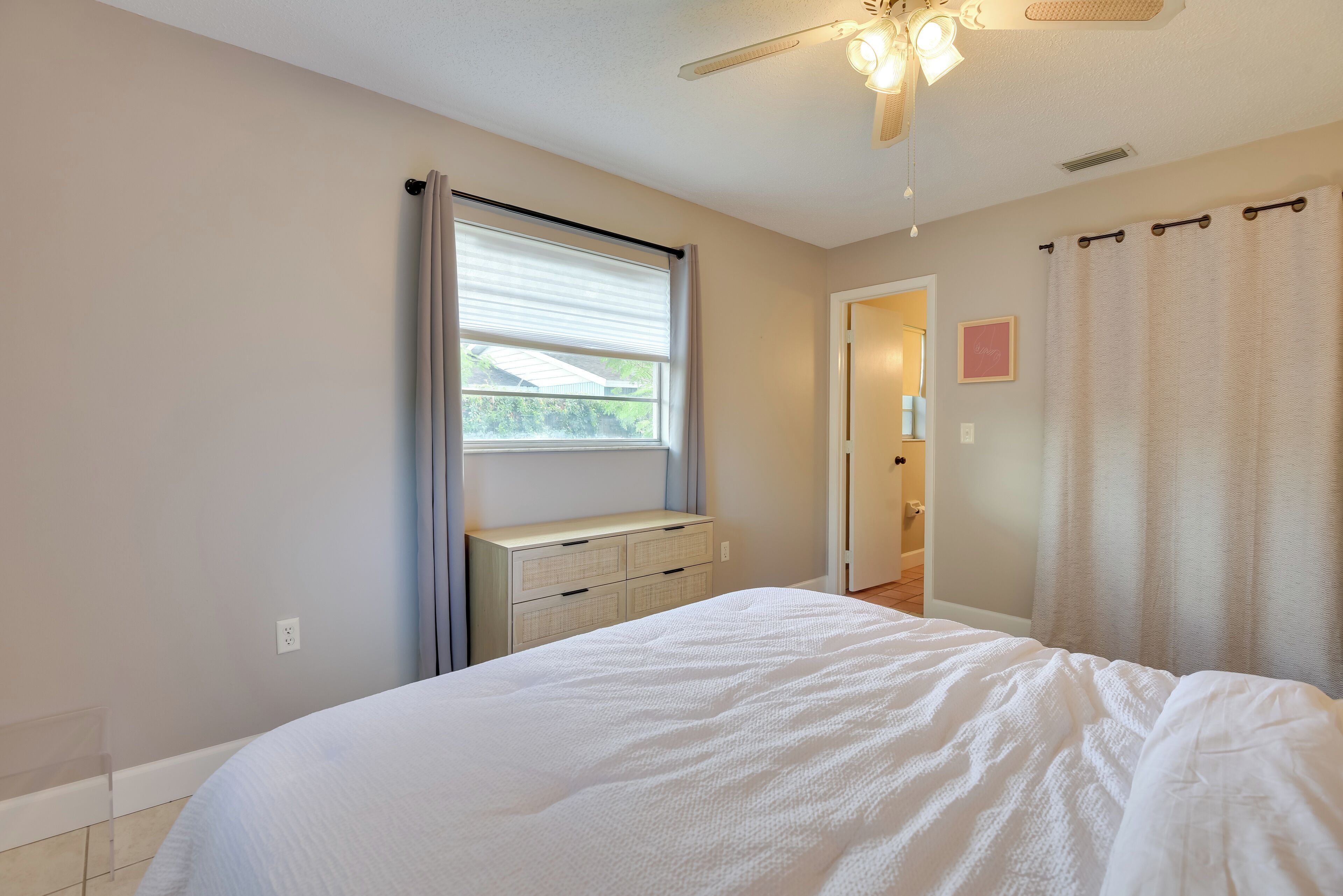 A bedroom with a bed, a ceiling fan, a window with curtains, a dresser, and a door leading to another room.