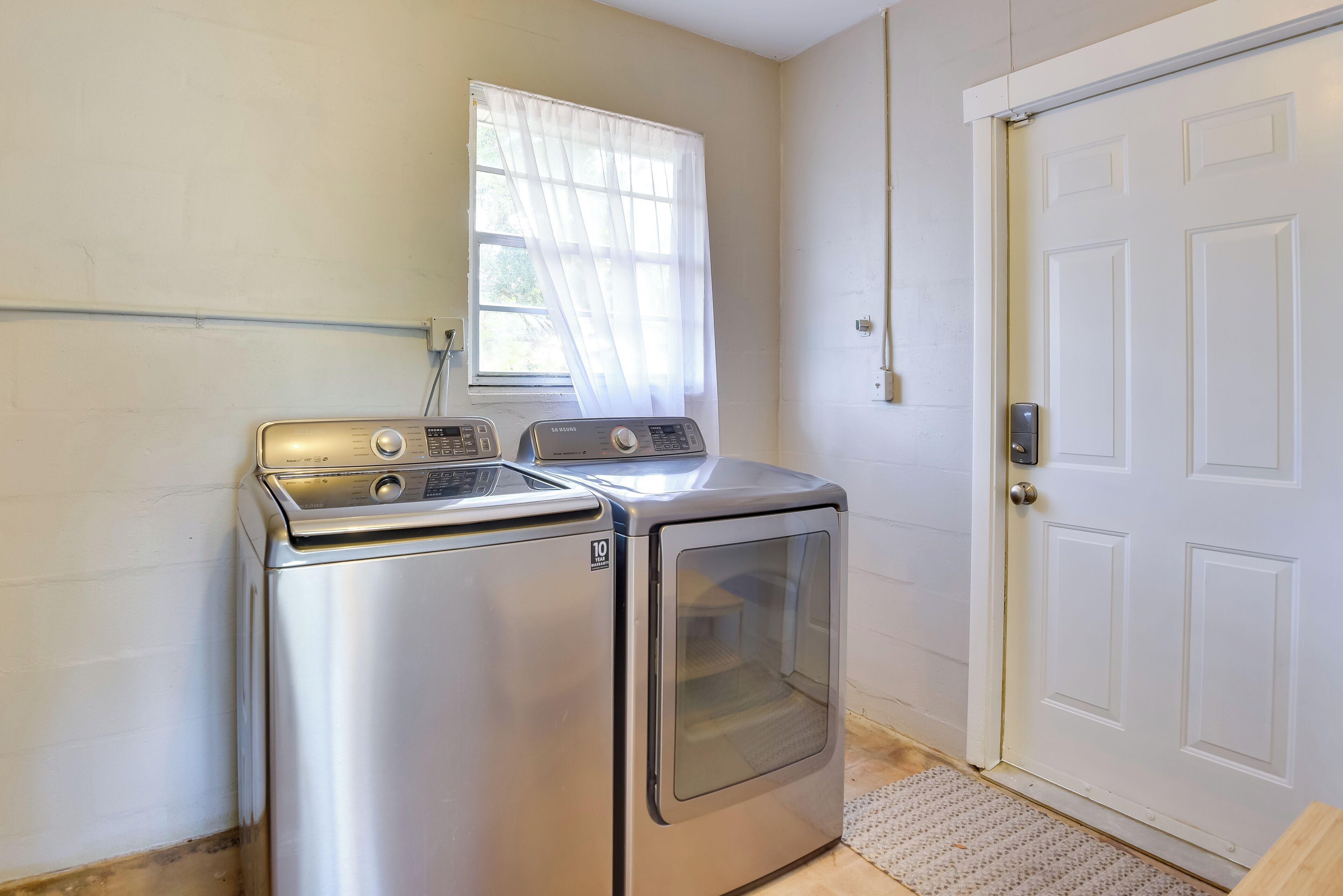 A laundry room with a front-loading washer and dryer, a window with sheer curtains, and a white door.