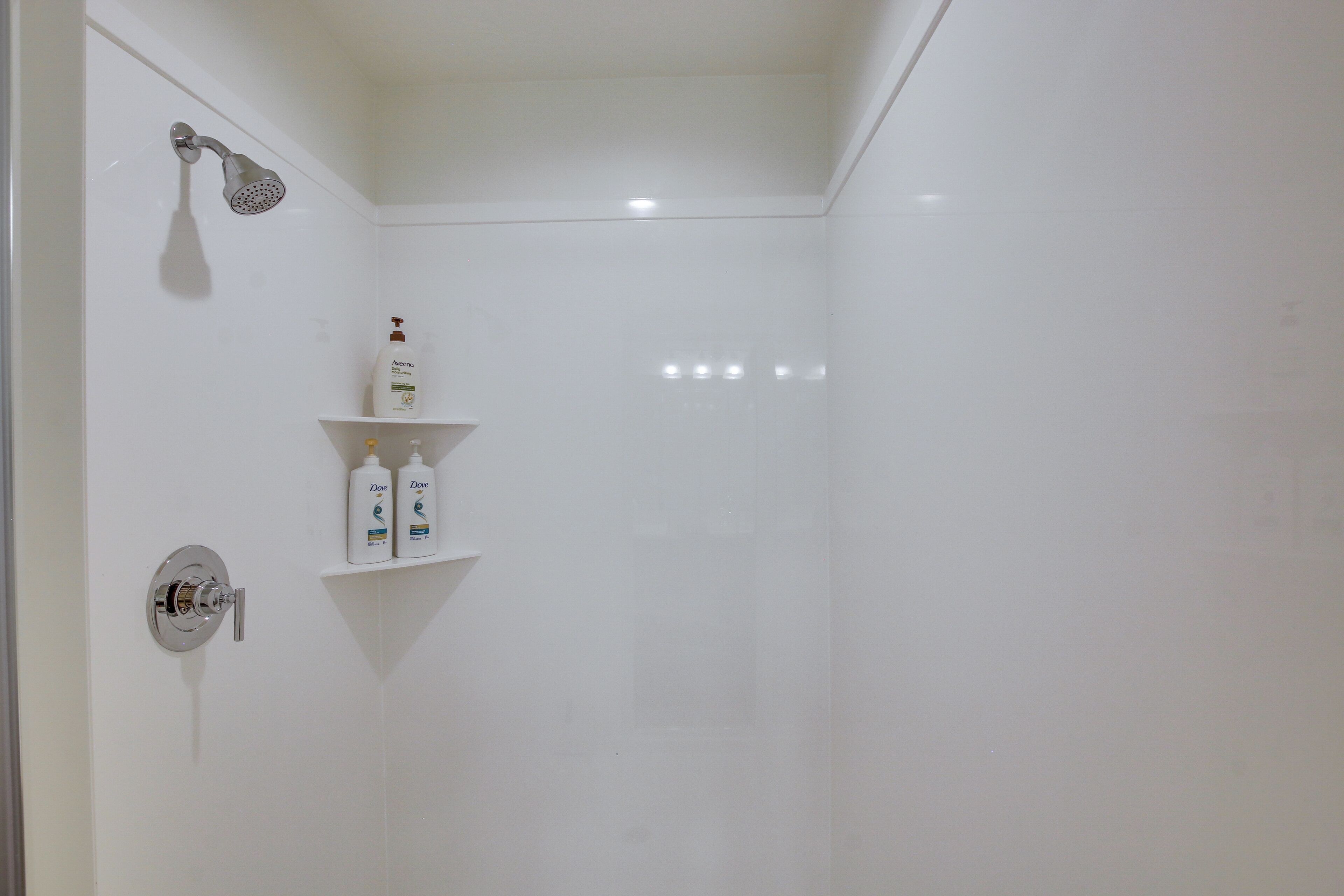 Eine weiße Dusche mit Duschbrause und Wandarmatur. Zwei weiße Ablagen mit Pflegeprodukten.