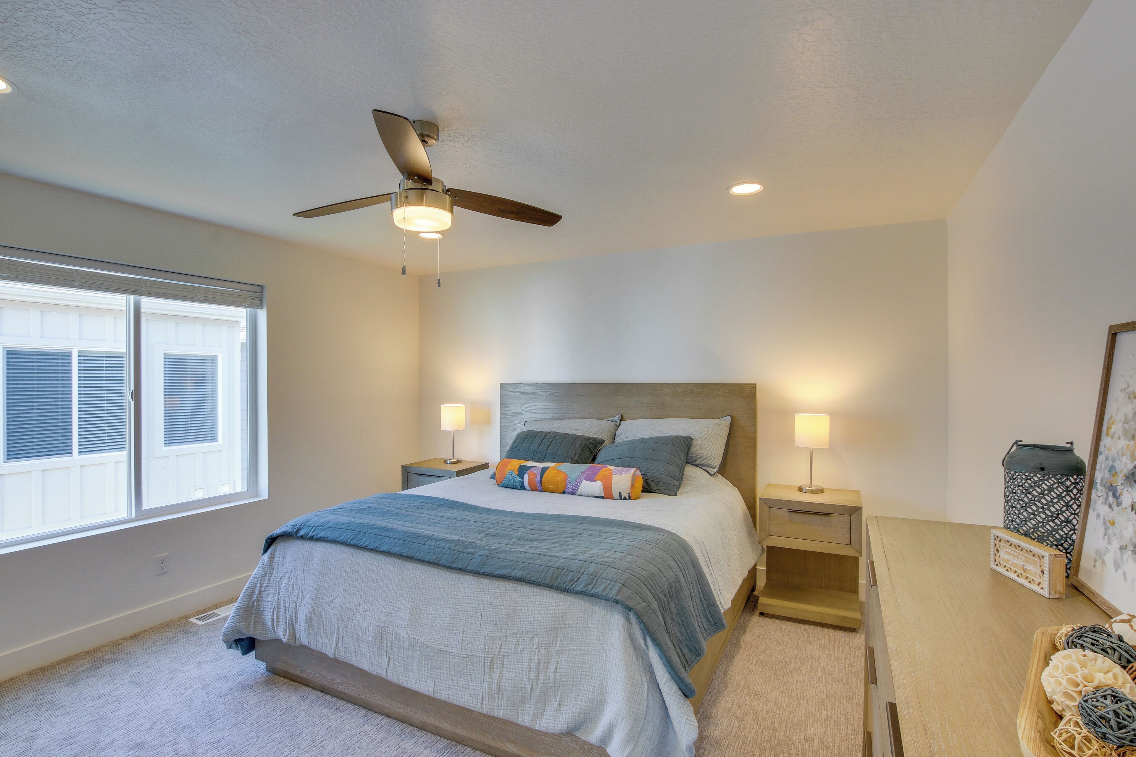 A bedroom with a bed, nightstands, a ceiling fan, a window with blinds, and a bedside table with a lamp.