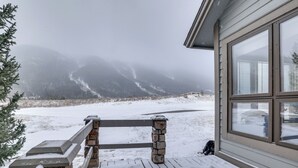 Ein verschneiter Balkon mit einer Stein Säule und einem Holzgeländer, mit Blick auf eine verschneite Landschaft und Berge.