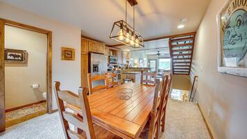 A spacious dining area with a wooden table and chairs, a hanging light fixture, and a kitchen in the background.