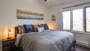 Un dormitorio con una cama grande, una mesita de noche con lámpara, un ventilador de techo y una ventana con vistas a árboles.