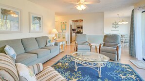 Olohuoneessa on harmaa sohva, raidallinen lepotuoli, lasinen sohvapöytä ja sininen matto. Huoneessa on myös kattotuuletin, lamppu sekä ruokailutila, jossa on tuoli ja pöytä.
