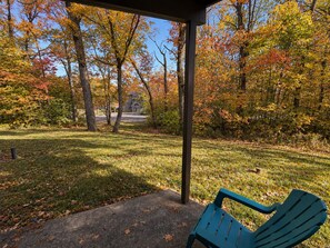 A covered outdoor seating area with a bench, surrounded by trees with autumn foliage, and a pathway leading to a building.