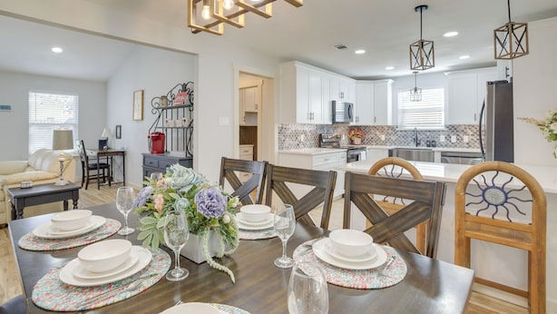 A dining area with a wooden table set for four, surrounded by chairs. A kitchen is visible in the background with white cabinets and a microwave. - Dog-friendly Home in The Woodlands w/ Fenced Yard! (Conroe)
