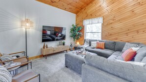 A modern living room with a grey sofa, a wooden ceiling, a flat-screen TV, a floor lamp, and a window with curtains. - Mtn-view Waynesville Home Near Trout Stocked Creek (Waynesville)