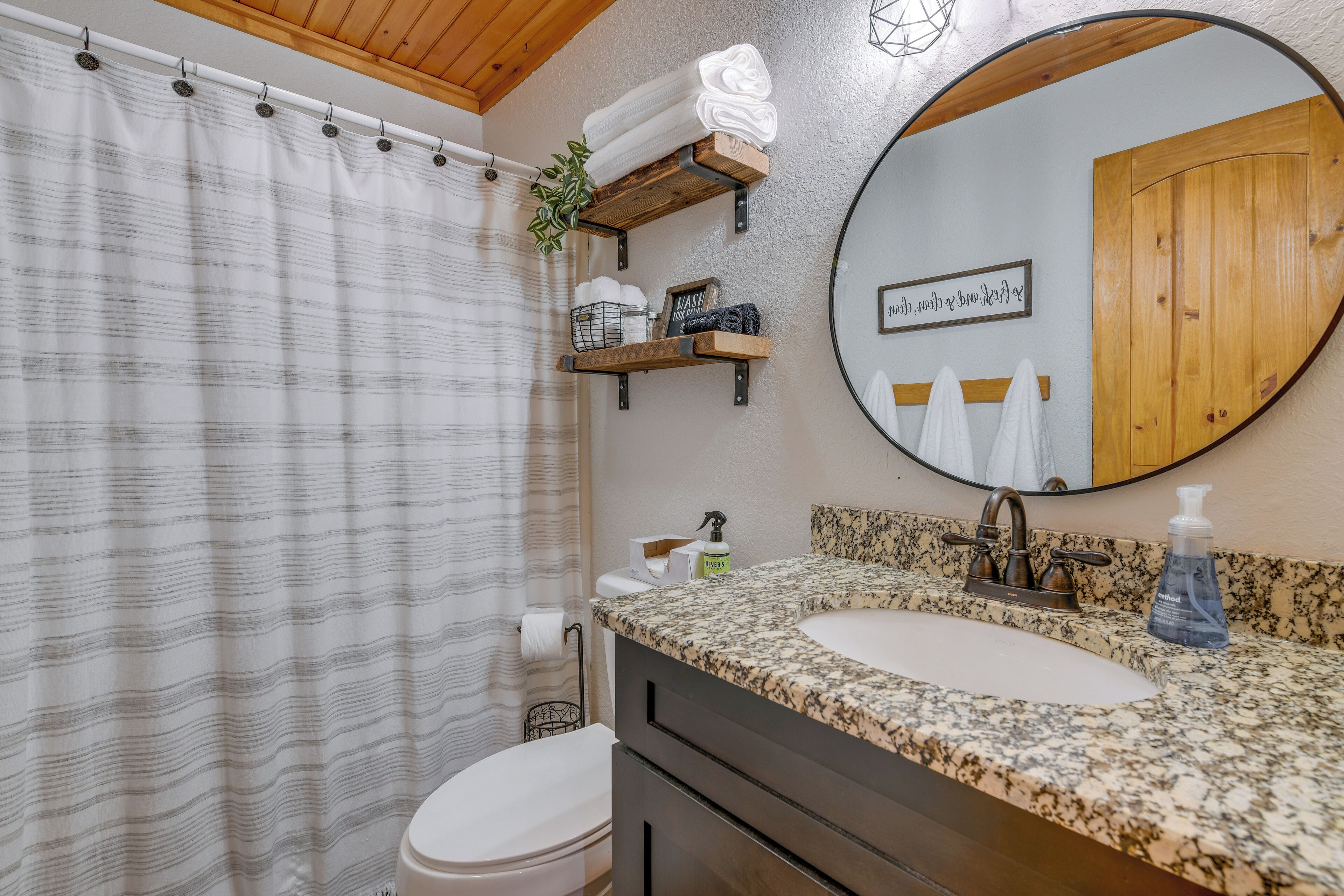 A bathroom with a striped shower curtain, a sink with a granite countertop, a wooden vanity, and a mirror with a wooden door reflection.