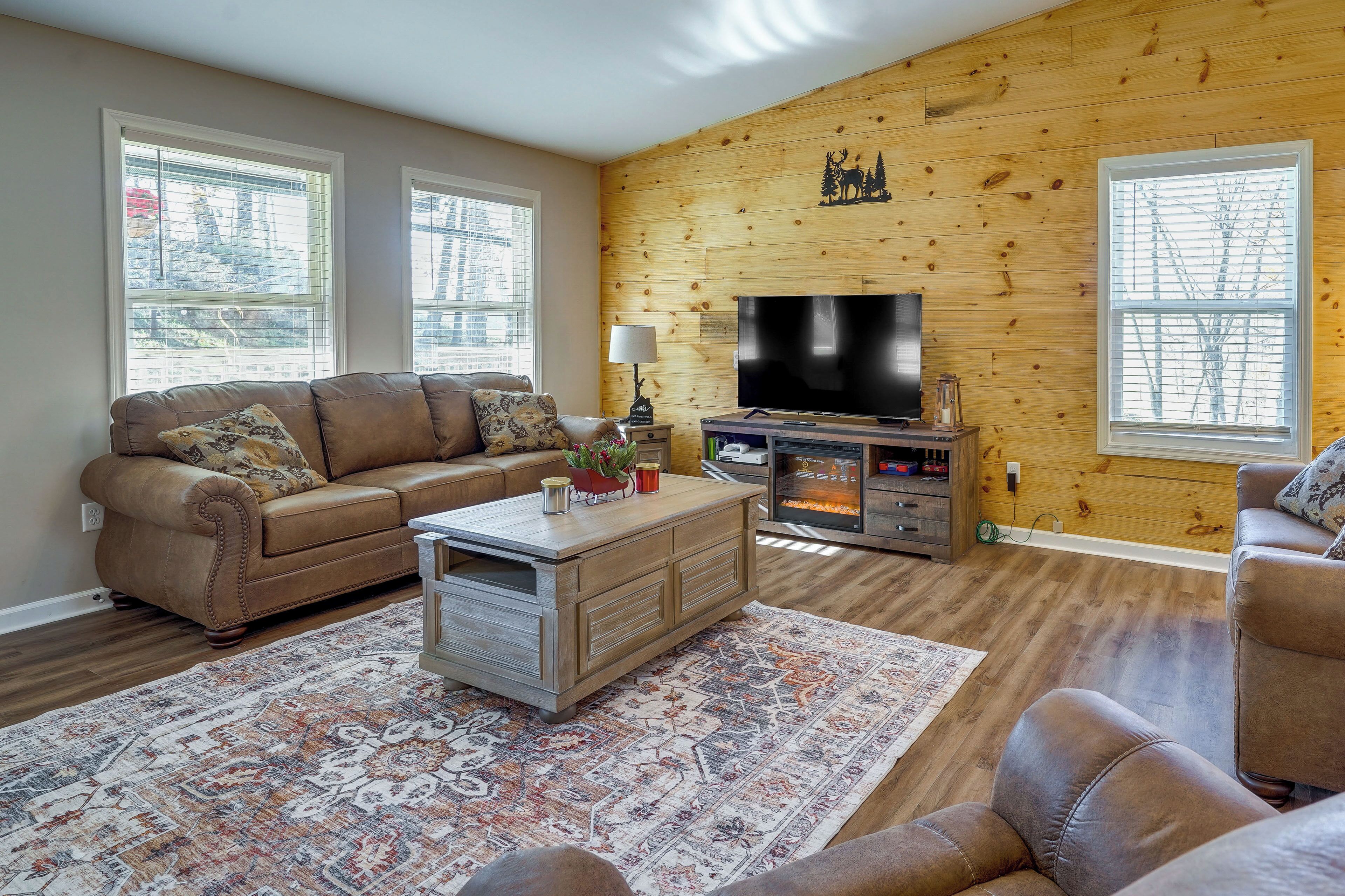 A living room with a brown leather sofa, a coffee table, a flat-screen TV, and a wooden wall with a deer and tree decoration.