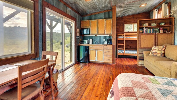 Interior rústico de cabana com mesa de jantar de madeira, sofá, área de cozinha e uma janela grande com vista para o exterior.