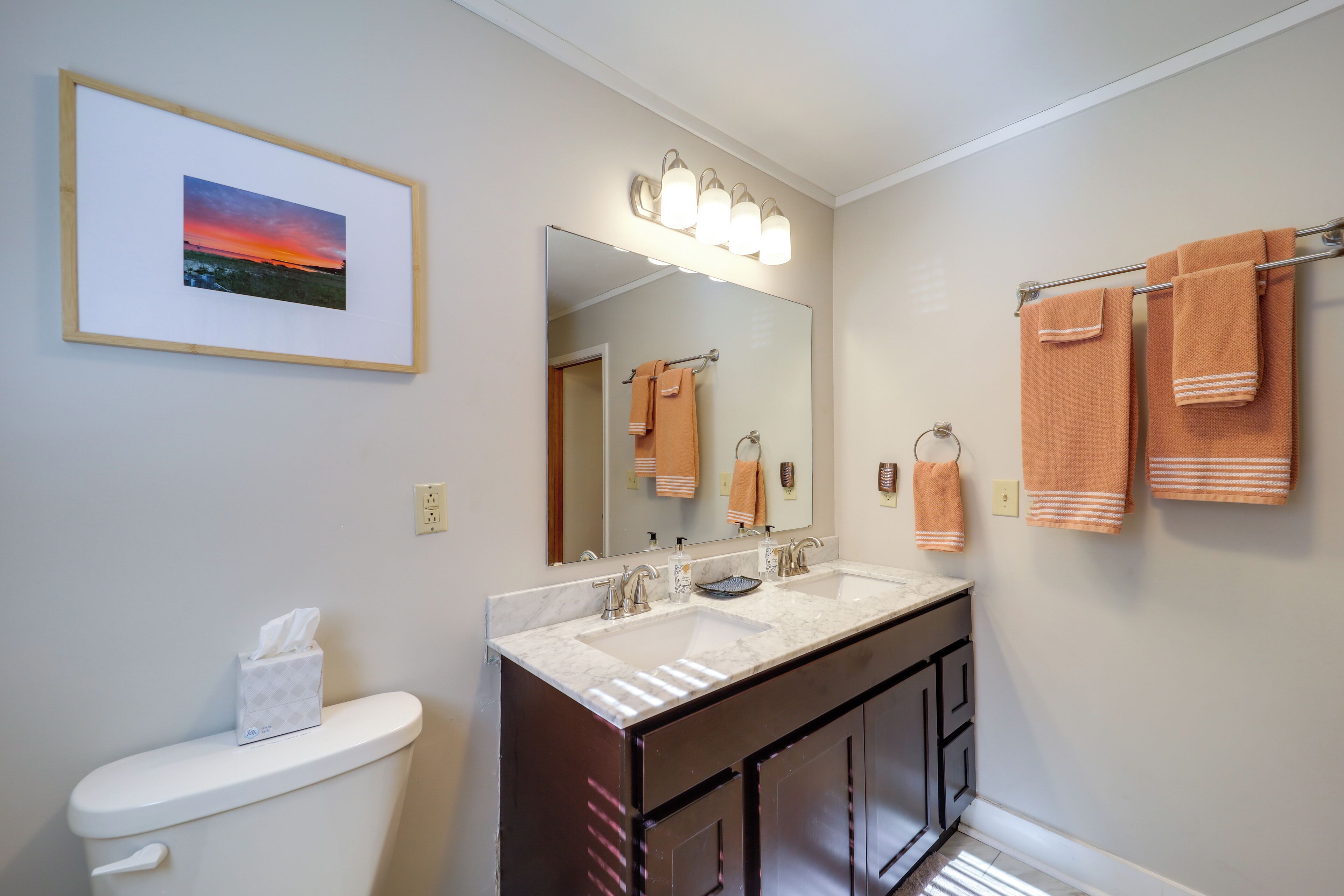 Casa de banho com uma pia dupla, sanita, um quadro emoldurado e toalhas penduradas num porta-toalhas.