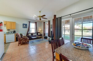 Amplia sala de estar con sofá, mesa de centro y una mesa de comedor con sillas. Se observa una zona de cocina con armarios y lavavajillas. Se ve un ventilador de techo, y la habitación tiene ventanas grandes con cortinas.
