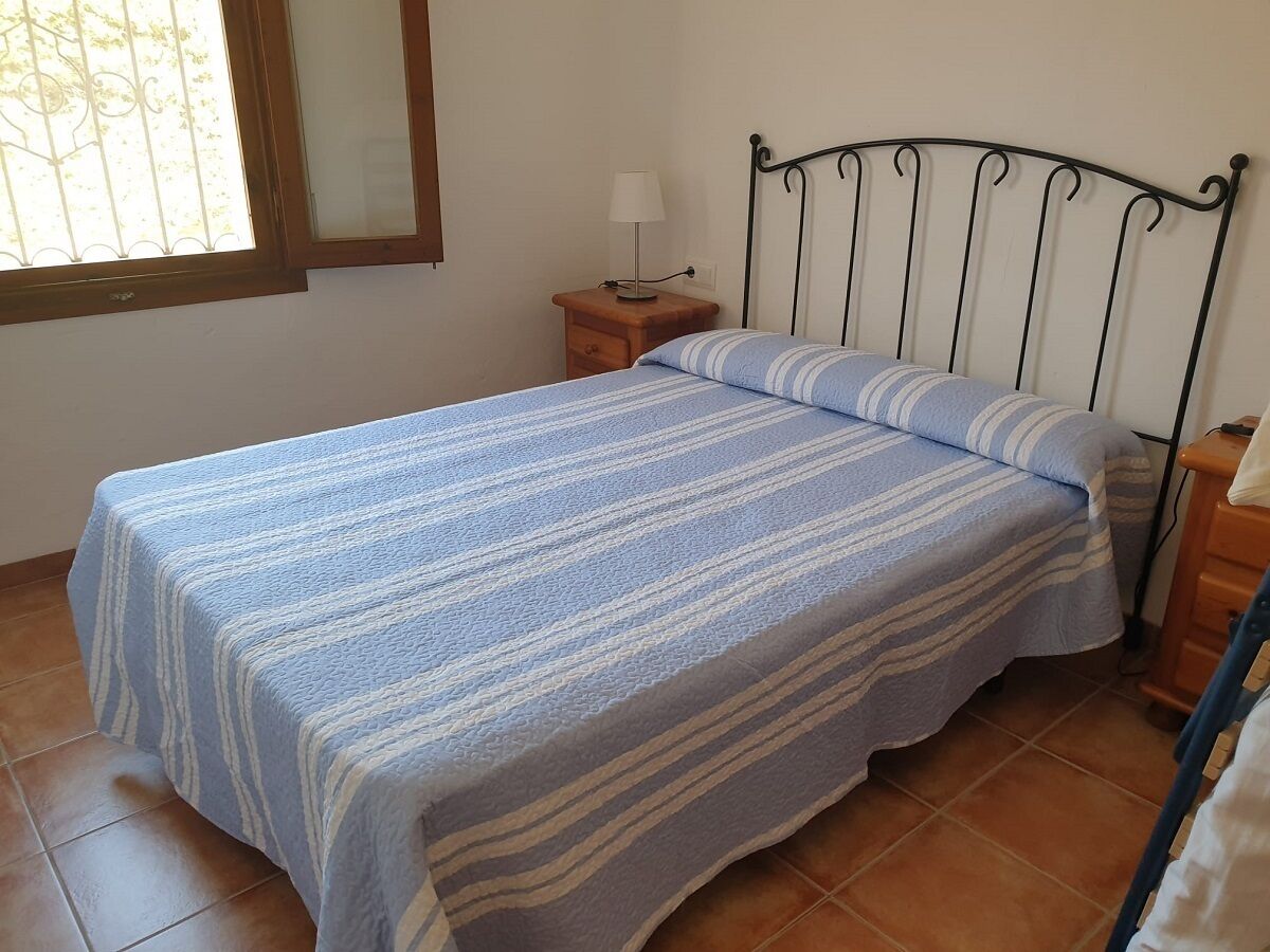 A bed with a striped bedspread, a nightstand with a lamp, and a window with bars.