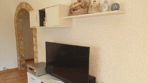 Una sala de estar con un televisor de pantalla plana sobre un mueble blanco, un estante pequeño con adornos encima, y un arco de piedra al fondo.