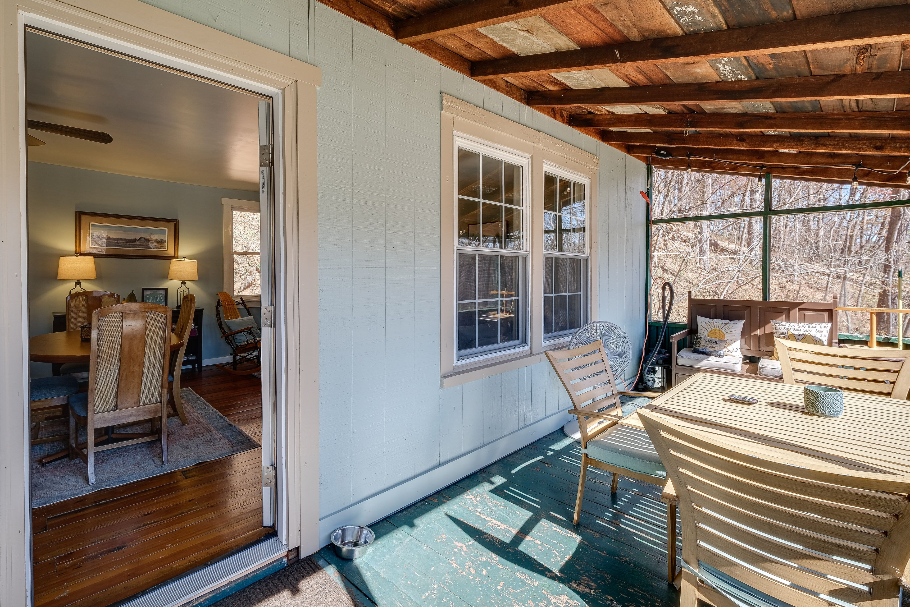 A cozy room with a wooden ceiling, a dining area with chairs and a table, and a window with a view of trees.