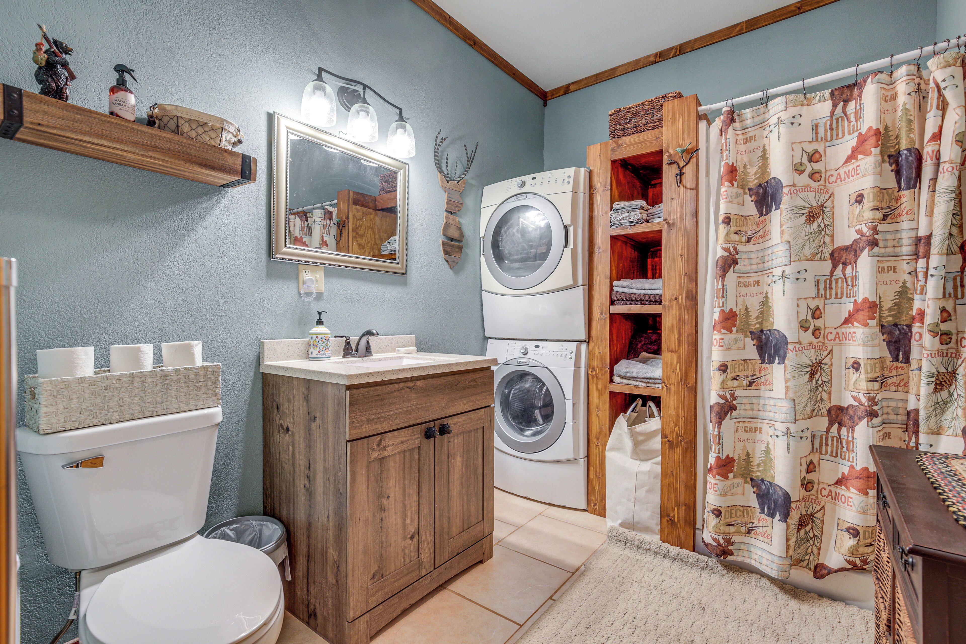 A compact bathroom with a toilet, sink, and washing machine, wooden cabinets, and a shower curtain with a nature theme.