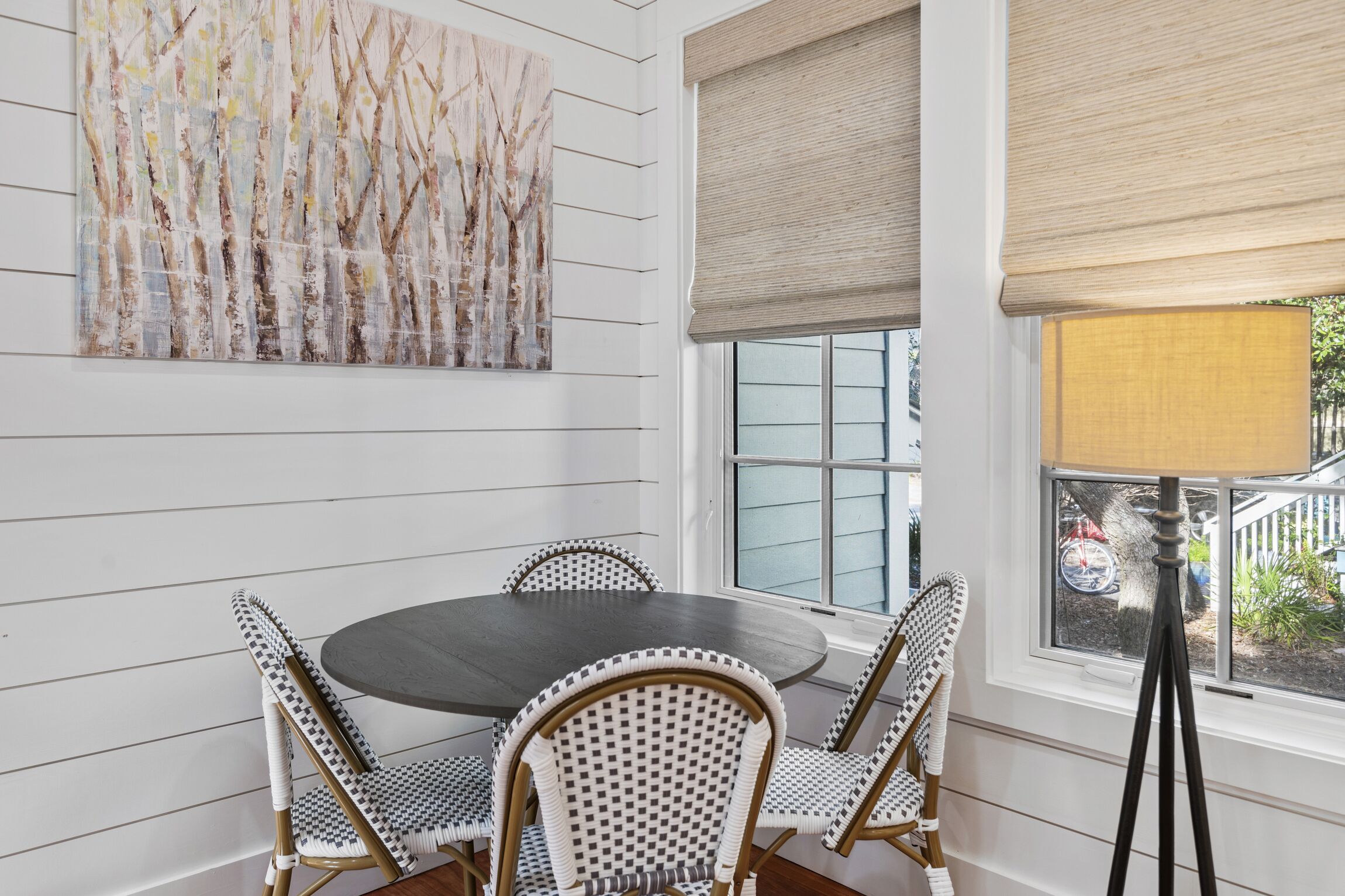A dining area with a round table and checkered chairs, a large abstract painting on the wall, and a window with a view of the outside.