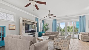 Un vaste salon doté d’un grand téléviseur à écran plat, de canapés beiges, d’une table basse et d’un pouf à motifs. On y trouve deux ventilateurs de plafond, une grande fenêtre offrant une vue sur la ville et un balcon avec des rideaux bleus.
