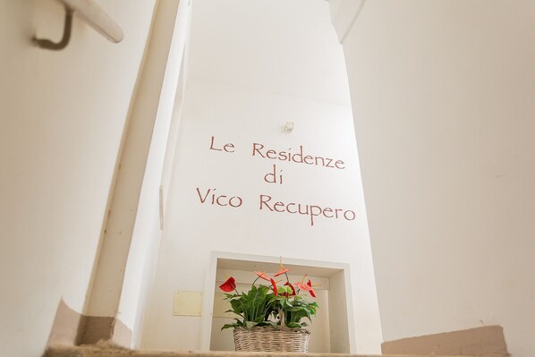 A white wall with a sign that reads 'Le Residenze di Vico Recupero' and a small potted plant with red flowers. - Residenza Vico Recupero by Wonderful Italy (Polignano a Mare)