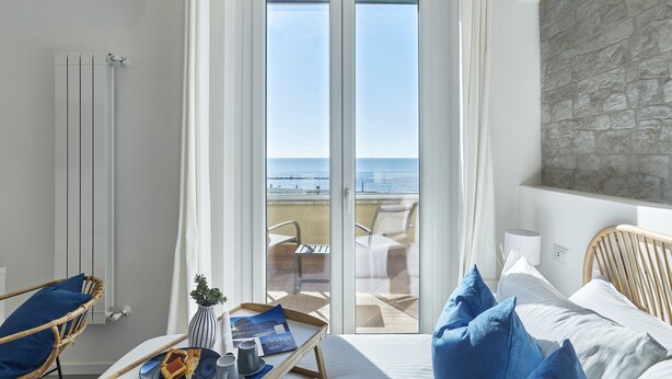 A bedroom with a bed, a wooden headboard, a bedside table with a lamp, a wicker chair, a view of the sea through a glass door, and a stone wall. - Charming penthouse with an ocean view a few steps from the train and bus station (Lido di Ostia)