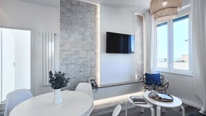 A modern room with a stone accent wall, a flat-screen TV, a dining area with a white table and chairs, and a window with a view. - Charming penthouse with an ocean view a few steps from the train and bus station (Lido di Ostia)