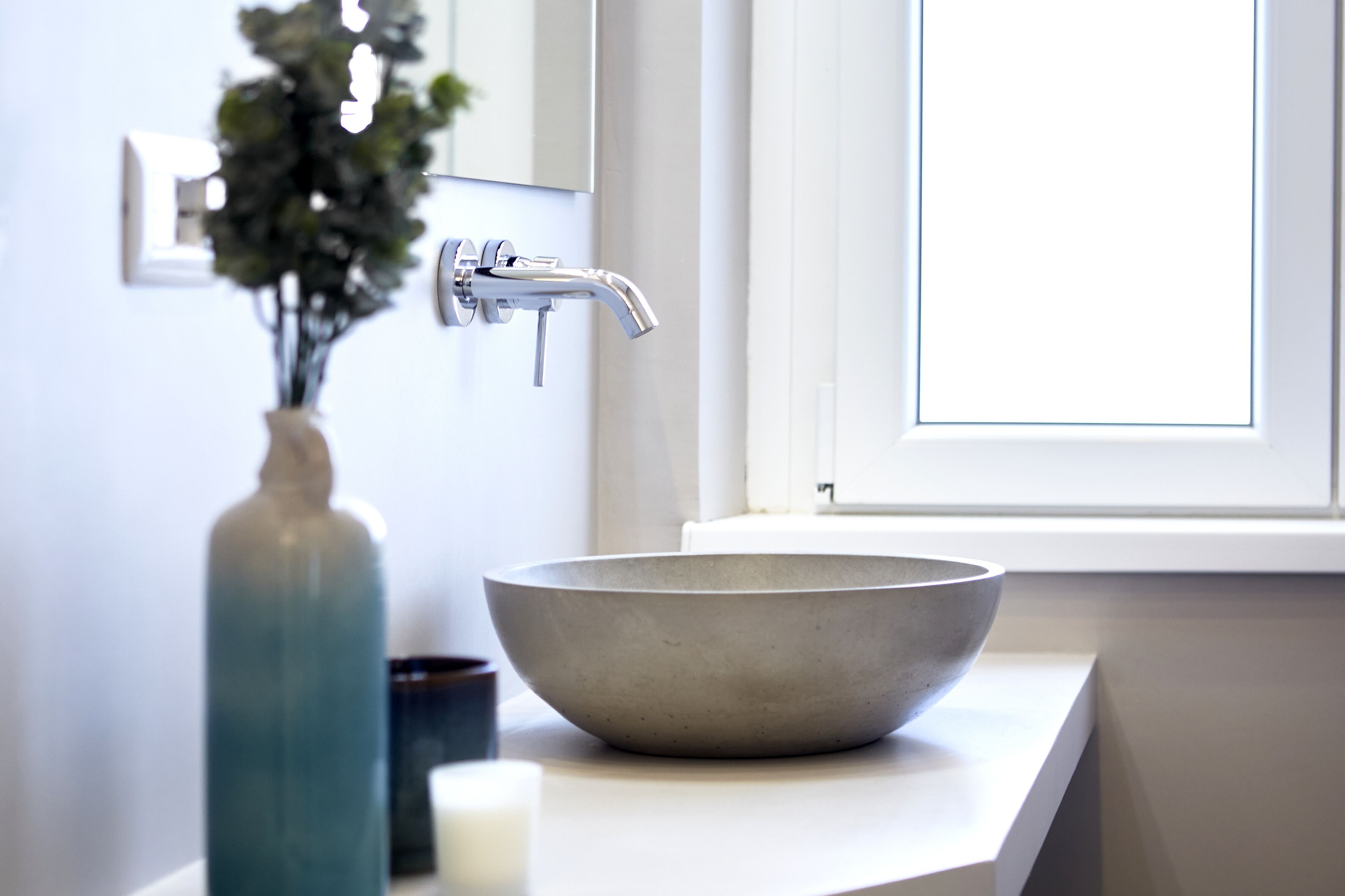 Een badkamer met een groot, rond, beige wastafel, een moderne kraan, een vaas met groene planten en een raam boven de wastafel.