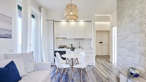 A modern living room with a white sofa, a dining table with chairs, and a kitchen area with white cabinets and a stone accent wall. - Charming penthouse with an ocean view a few steps from the train and bus station (Lido di Ostia)