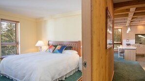 Quarto com cama grande, cabeceira de madeira, mesa de cabeceira com abajur e vista para a cozinha.