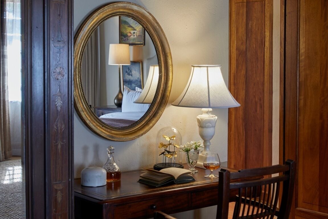 A wooden desk with a mirror, a lamp, a book, a glass, and a decorative item.
