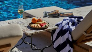 Escena junto a la piscina con una mesa que sostiene una copa de vino blanco, un plato de frutas cortadas y una revista.