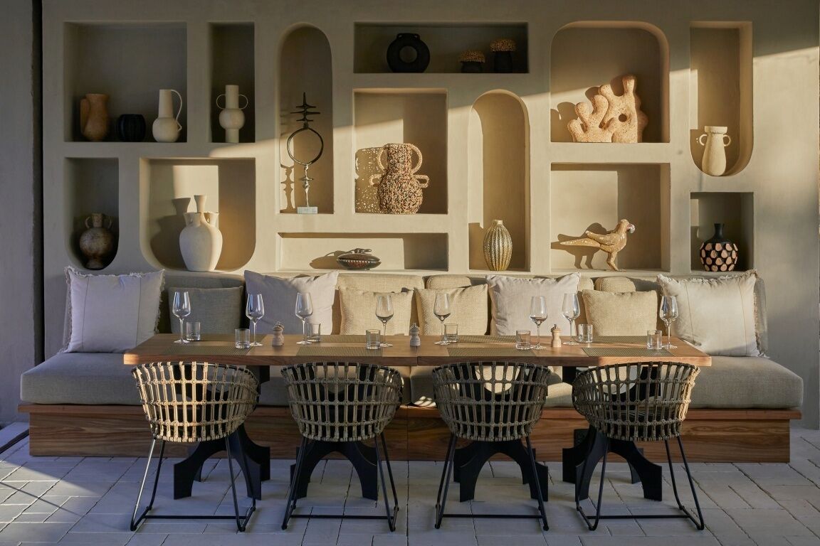 Un comedor con una mesa de madera y sillas de mimbre, un sofá con cojines y estantes con diversos objetos decorativos.
