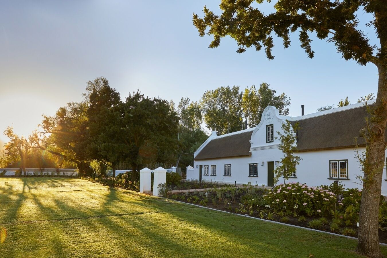 A white colonial-style house with a thatched roof, surrounded by lush greenery and flowering plants.