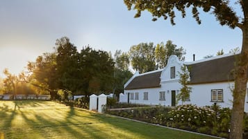 A white colonial-style house with a thatched roof, surrounded by lush greenery and flowering plants.
