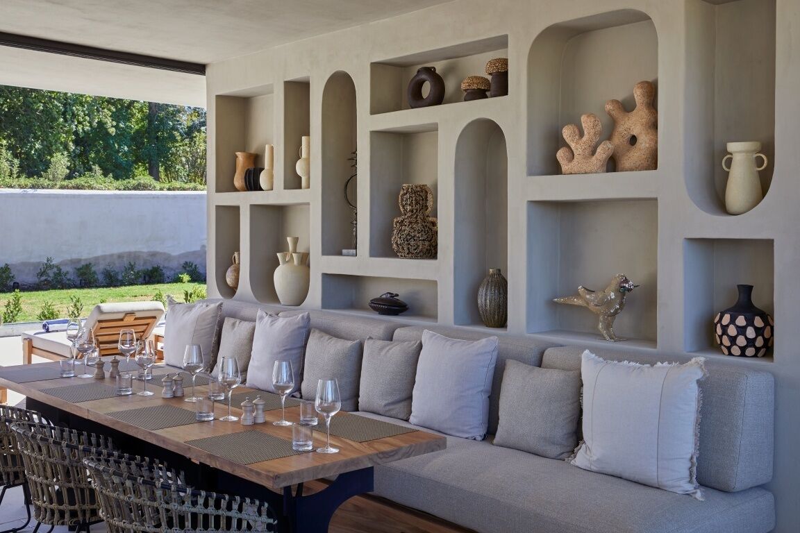 Un comedor al aire libre moderno con una mesa de madera dispuesta para seis personas, rodeado por un sofá gris con cojines.