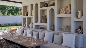 Un espace repas extérieur moderne, avec une table en bois disposée pour accueillir six personnes, entourée d’un canapé gris agrémenté de coussins.