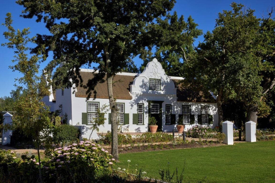 A white building with green shutters, surrounded by a well-maintained garden and trees.