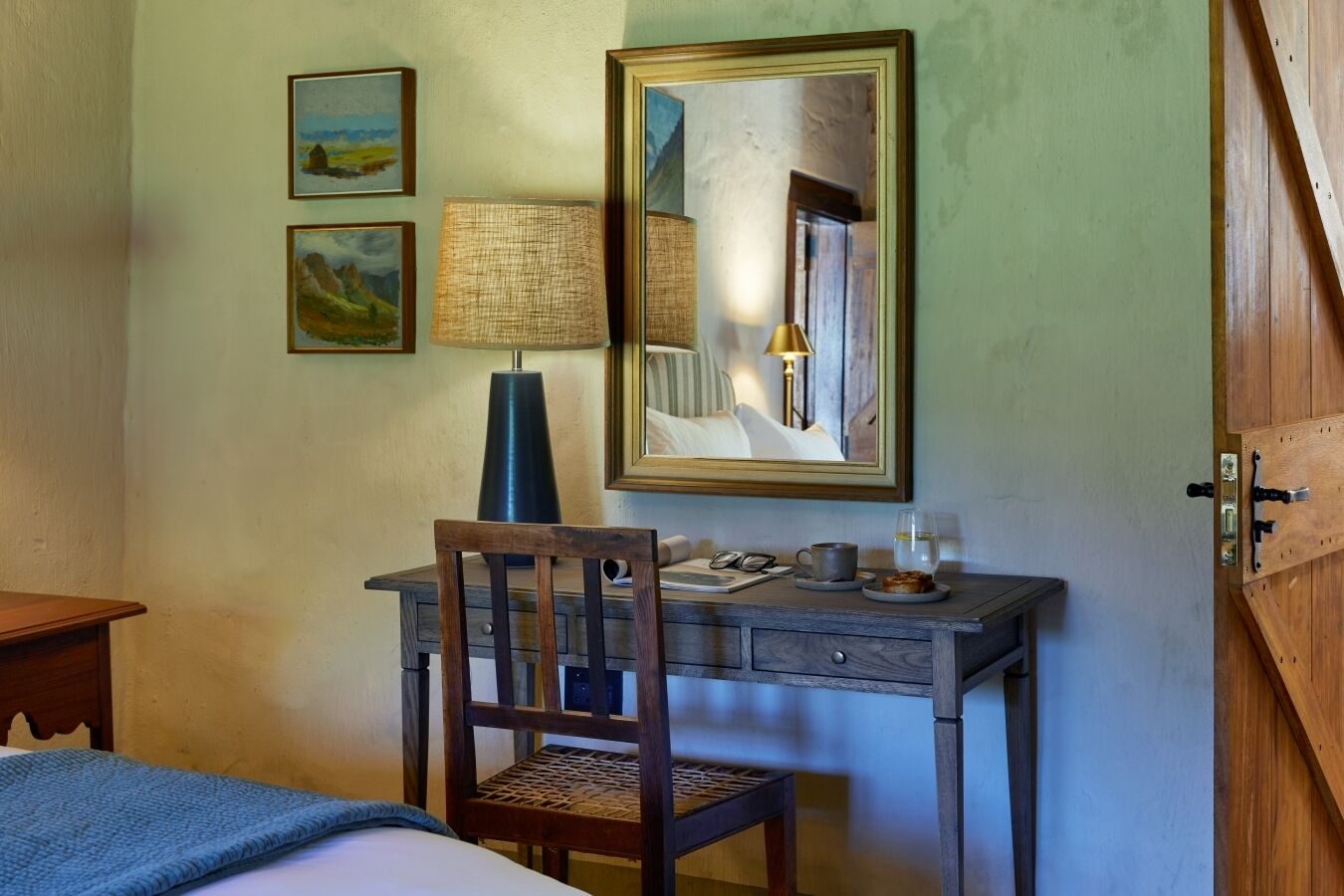 A room with a wooden desk, a chair, a bed, and a wardrobe. There are framed pictures on the wall and a lamp on the desk.