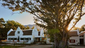 Un edificio de estilo colonial blanco con un árbol grande en su frente, rodeado por un camino de grava y follaje.
