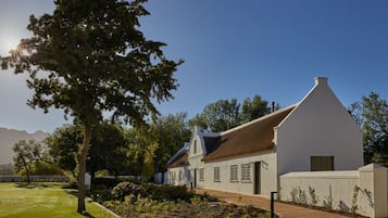 A white colonial-style building with a steeply pitched roof, surrounded by a well-maintained garden and a clear sky.