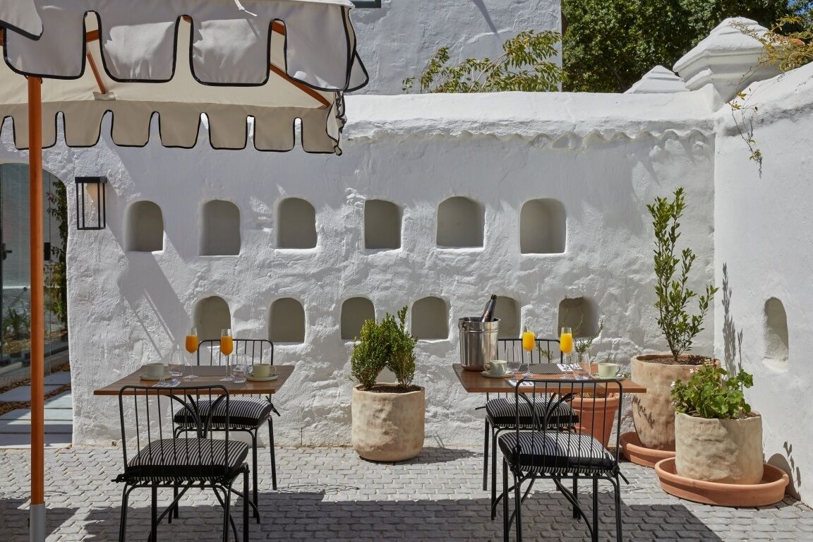 An outdoor seating area with tables and chairs, potted plants, and a white wall with arched windows.