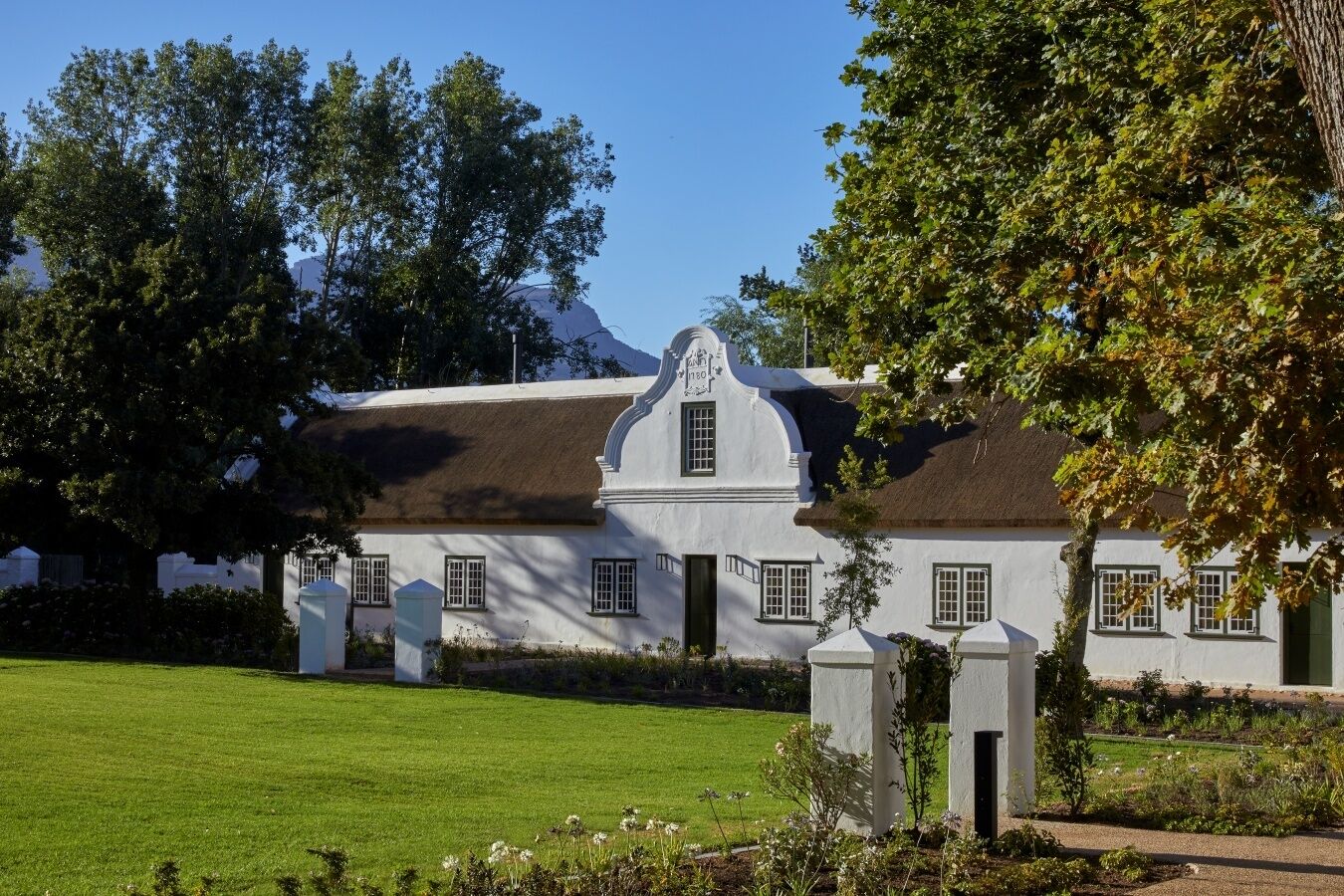 Un bâtiment blanc avec un toit de chaume, entouré d’une pelouse bien entretenue et d’arbres.