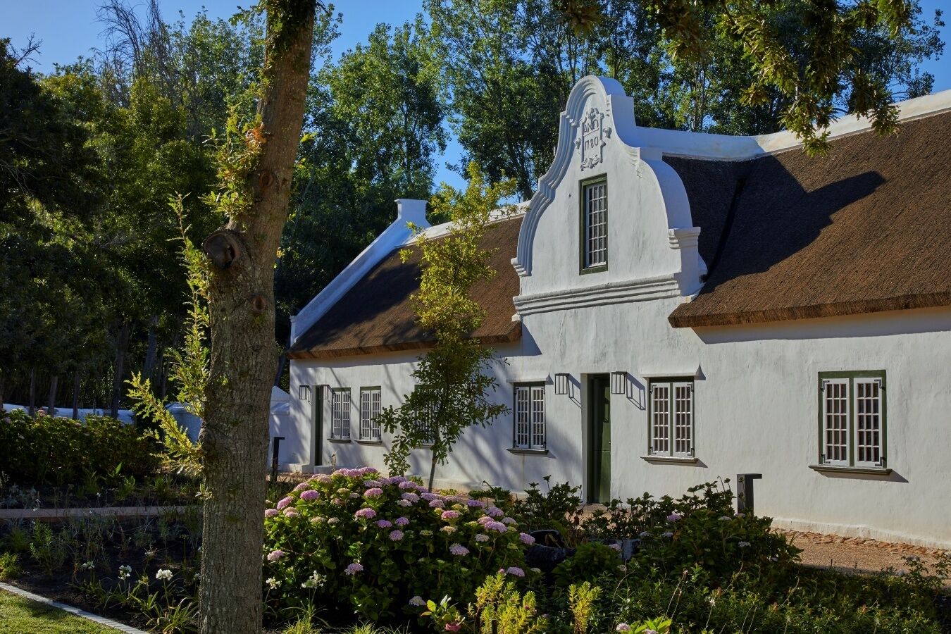 A white building with a thatched roof, surrounded by lush greenery and flowering plants.