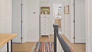 Un couloir avec une table en bois, un tapis coloré, une commode blanche avec des tiroirs et une porte donnant sur une salle de bain équipée d’un lavabo et d’un miroir.