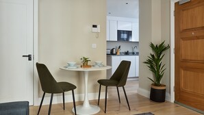 Une cuisine moderne avec des armoires blanches, un four à micro-ondes et un évier. On y trouve un coin repas avec une table blanche et deux chaises. Une plante en pot est visible à côté de la porte.