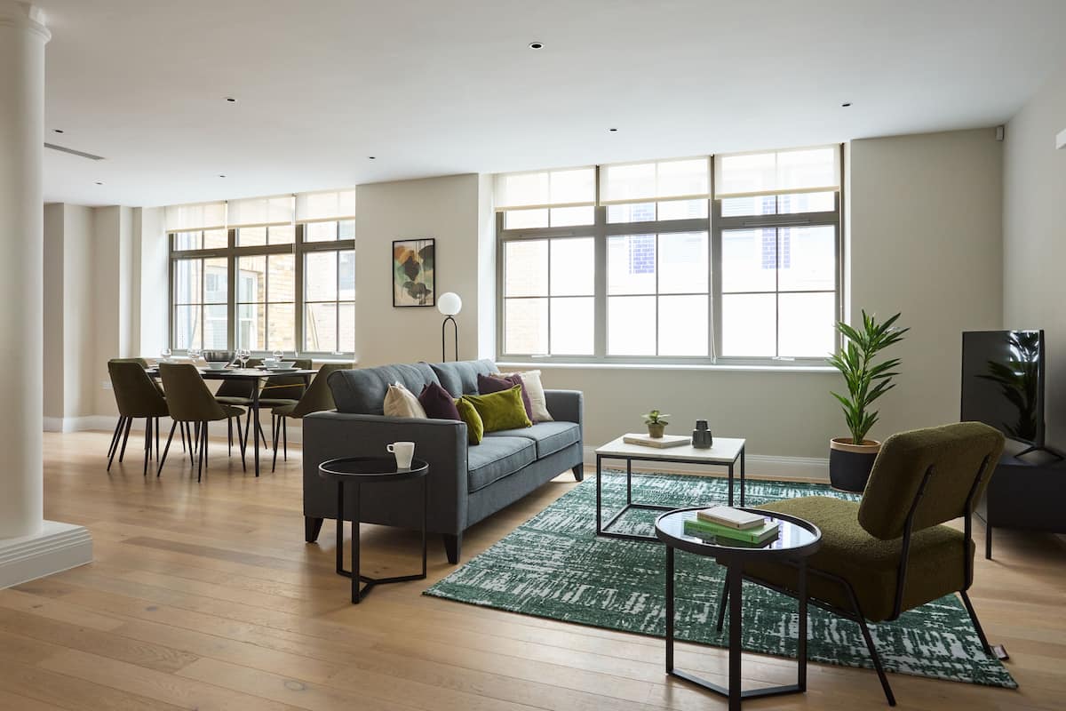 A spacious living room with a grey sofa, dining table, and chairs. There are large windows providing natural light and a view of the outside.
