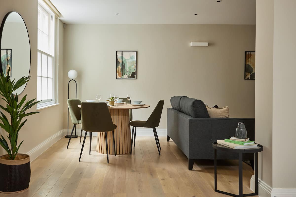 A modern living room with a round wooden table, two chairs, a grey sofa, and a small side table with books.