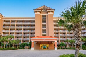 A multi-story hotel with a central entrance, balconies, and palm trees.