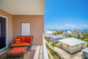 Un balcon avec un canapé et un pouf en osier, une porte blanche et une vue sur les maisons de plage.